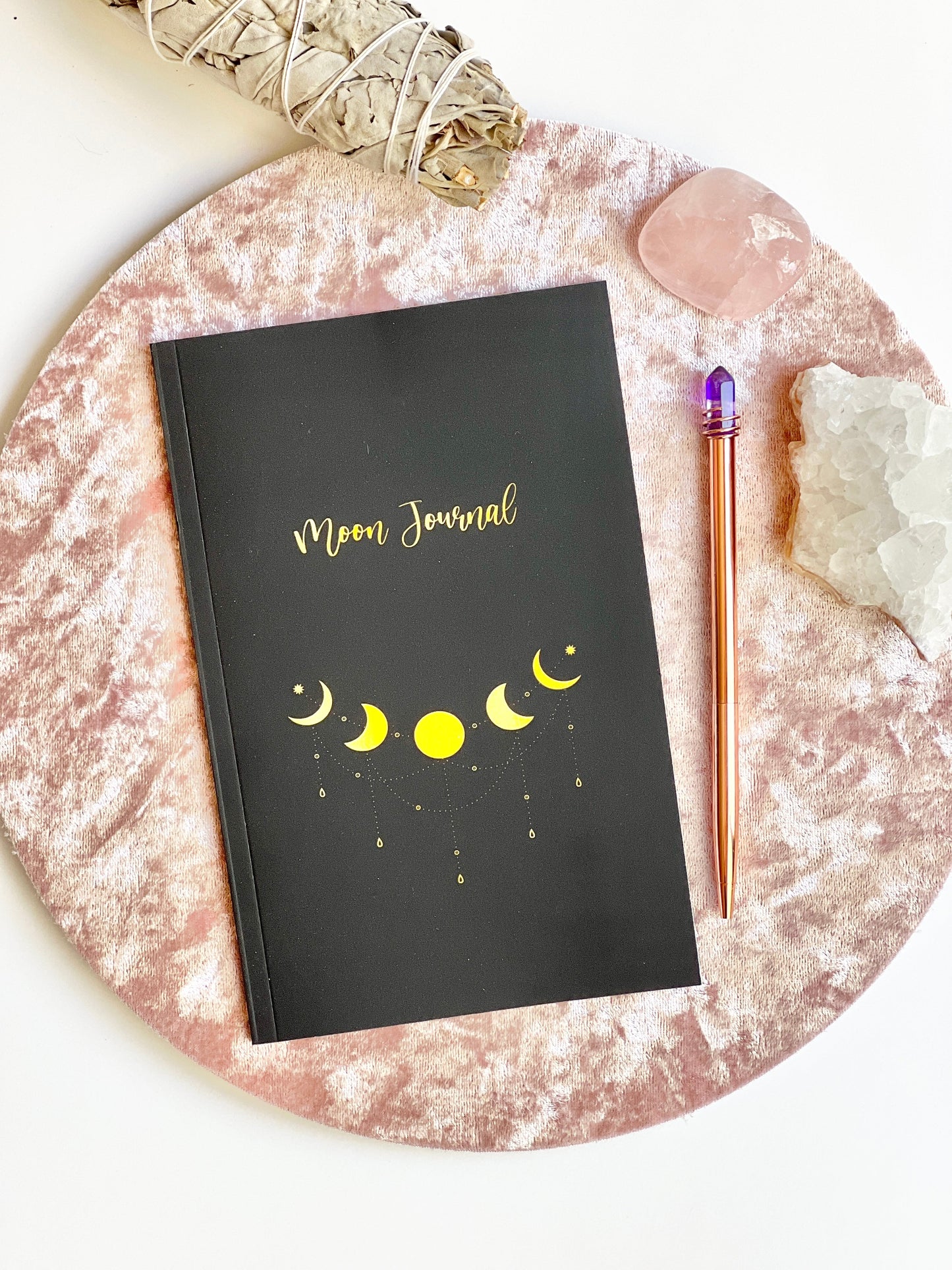 Front cover of Moon Rituals & Manifestation Journal in black glossy softcover with gold moon phase design displayed on a blush velvet surface with sage and crystals — for Carpe Diem Dream