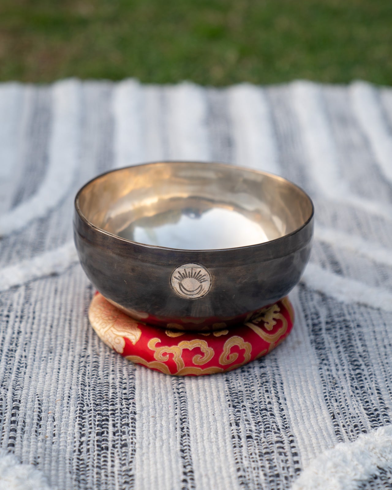 Tibetan Root Chakra New Moon singing bowl with engraved sun and moon symbol resting on a red patterned cushion on a textured woven blanket background. — for Carpe Diem Dream