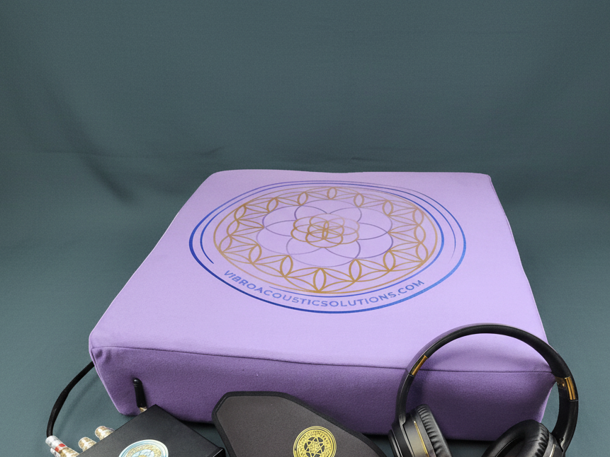 A vibroacoustic therapy zafu meditation cushion bundle featuring a lavender cushion with a gold lotus mandala, a compact black amplifier, over-ear headphones, and a satin eye mask arranged on a muted teal studio background. — for Carpe Diem Dream