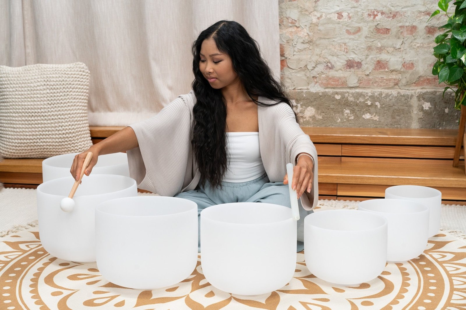 Woman seated on patterned rug playing seven white frosted crystal singing bowls in 14 inch through 8 inch sizes during a sound bath session — for Carpe Diem Dream