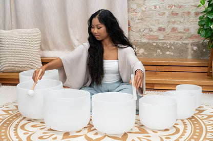 Woman seated on patterned rug playing seven white frosted crystal singing bowls in 14 inch through 8 inch sizes during a sound bath session — for Carpe Diem Dream
