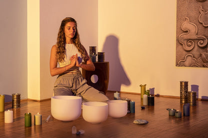 Woman seated on wooden floor with three white frosted crystal singing bowls in 12 inch, 10 inch, and 8 inch sizes surrounded by candles in a softly lit room — for Carpe Diem Dream