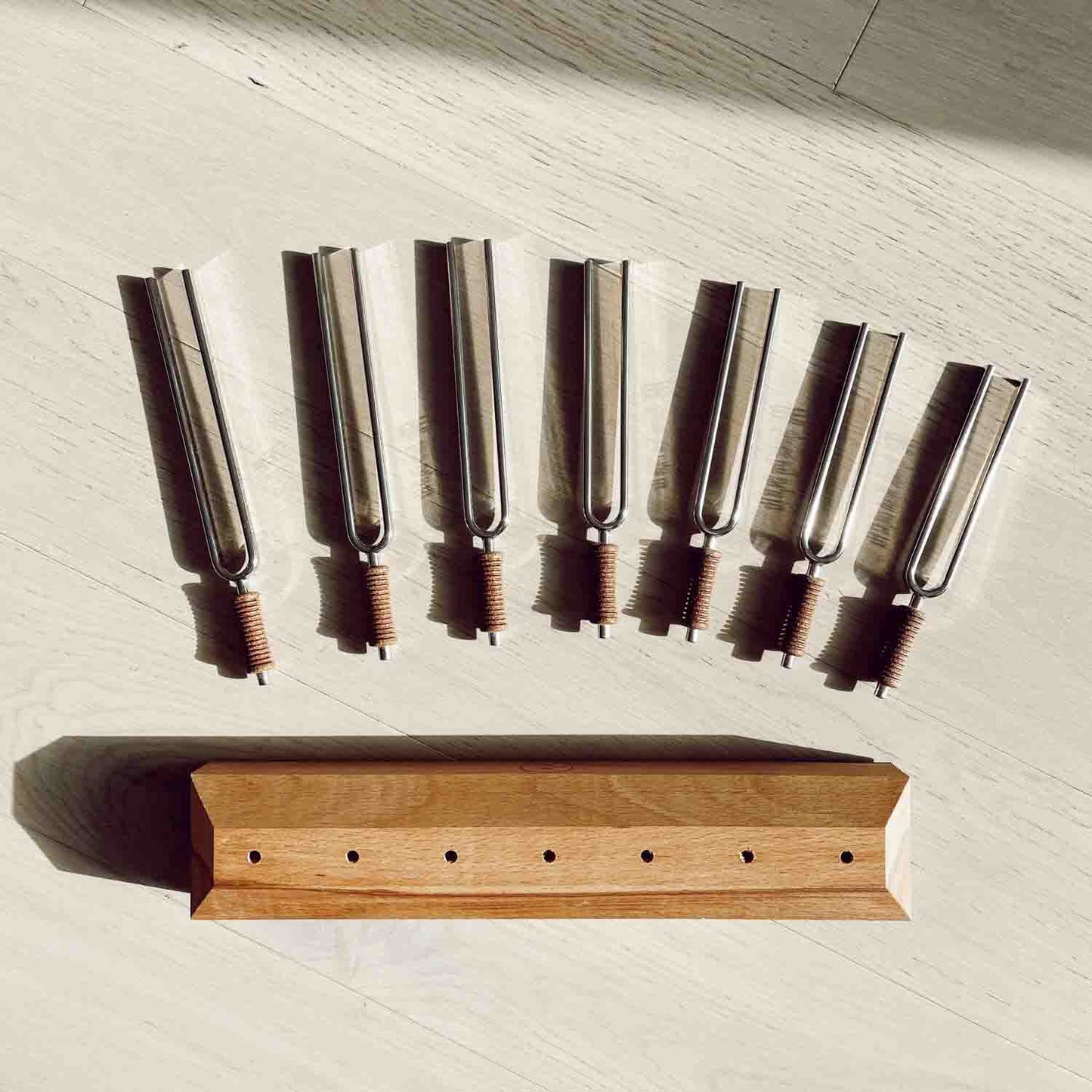 Seven chakra tuning forks with cork handles arranged above a wooden storage base in a top-down view on a light surface. — for Carpe Diem Dream