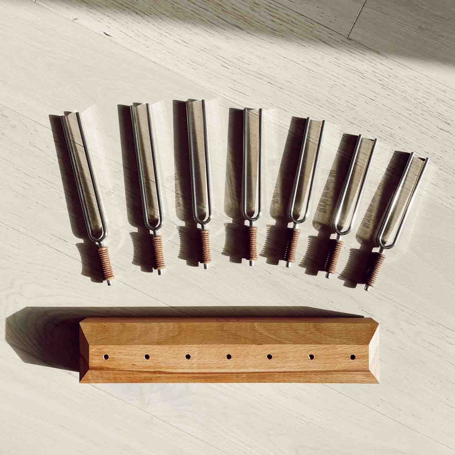 Seven chakra tuning forks with cork handles arranged above a wooden storage base in a top-down view on a light surface. — for Carpe Diem Dream