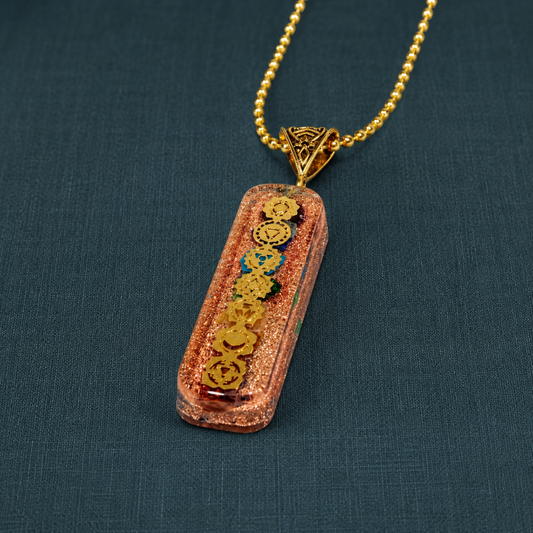 7 chakra orgone copper pendant with gold-tone chain, colorful chakra symbols, resin finish, copper accents, and dark teal linen backdrop. — for Carpe Diem Dream