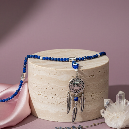 Silver dream catcher necklace with 999 angel number pendant and blue gemstone bead chain draped over stone pedestal beside clear quartz on soft mauve backdrop. — for Carpe Diem Dream
