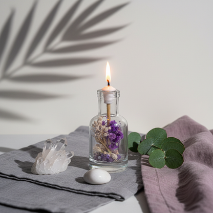 A refillable Aquarius oil candle in a clear glass vessel with dried purple and neutral flowers, lit on a soft linen surface with a crystal cluster, smooth stone, eucalyptus stems, and botanical shadows on a light wall. — for Carpe Diem Dream