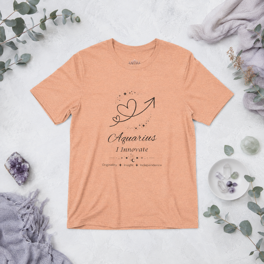 Aquarius Rising I Innovate zodiac T-shirt in Heather Peach with black celestial heart and arrow artwork and Originality, Insight, Independence text, styled in a cool-toned studio botanical flat lay. — for Carpe Diem Dream