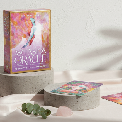 Ascension Oracle Deck displayed on gray travertine cylinders over blush silk fabric with cards laid flat, styled with eucalyptus sprig, rose quartz crystal, and soft daylight shadows by Carpe Diem Dream.