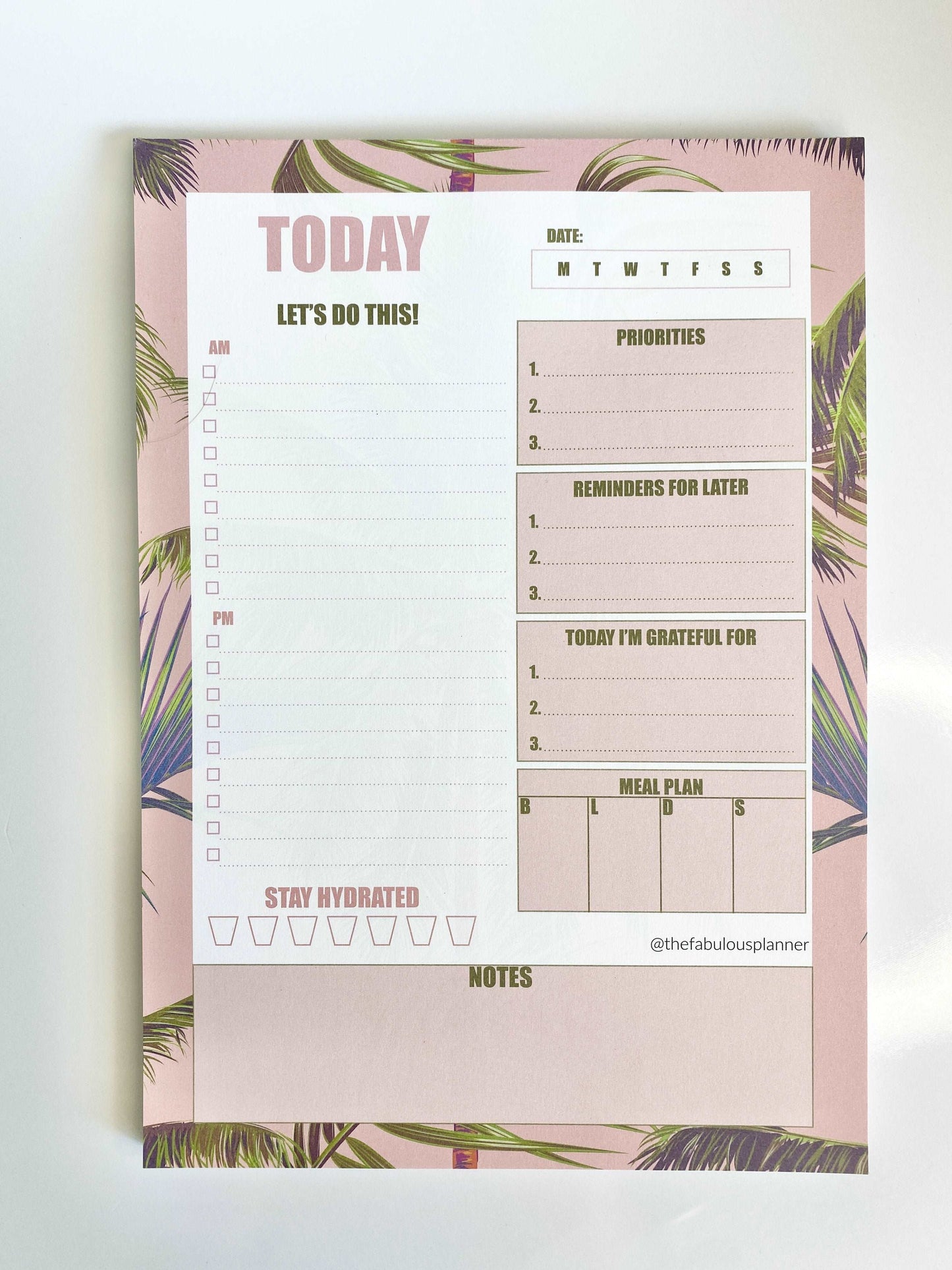 Close-up view of pink tropical leaves 7x10 daily planner notepad showing priorities, reminders, gratitude, meal plan, and water tracker sections on a white background — for Carpe Diem Dream
