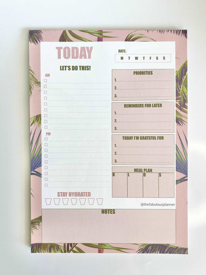 Close-up view of pink tropical leaves 7x10 daily planner notepad showing priorities, reminders, gratitude, meal plan, and water tracker sections on a white background — for Carpe Diem Dream