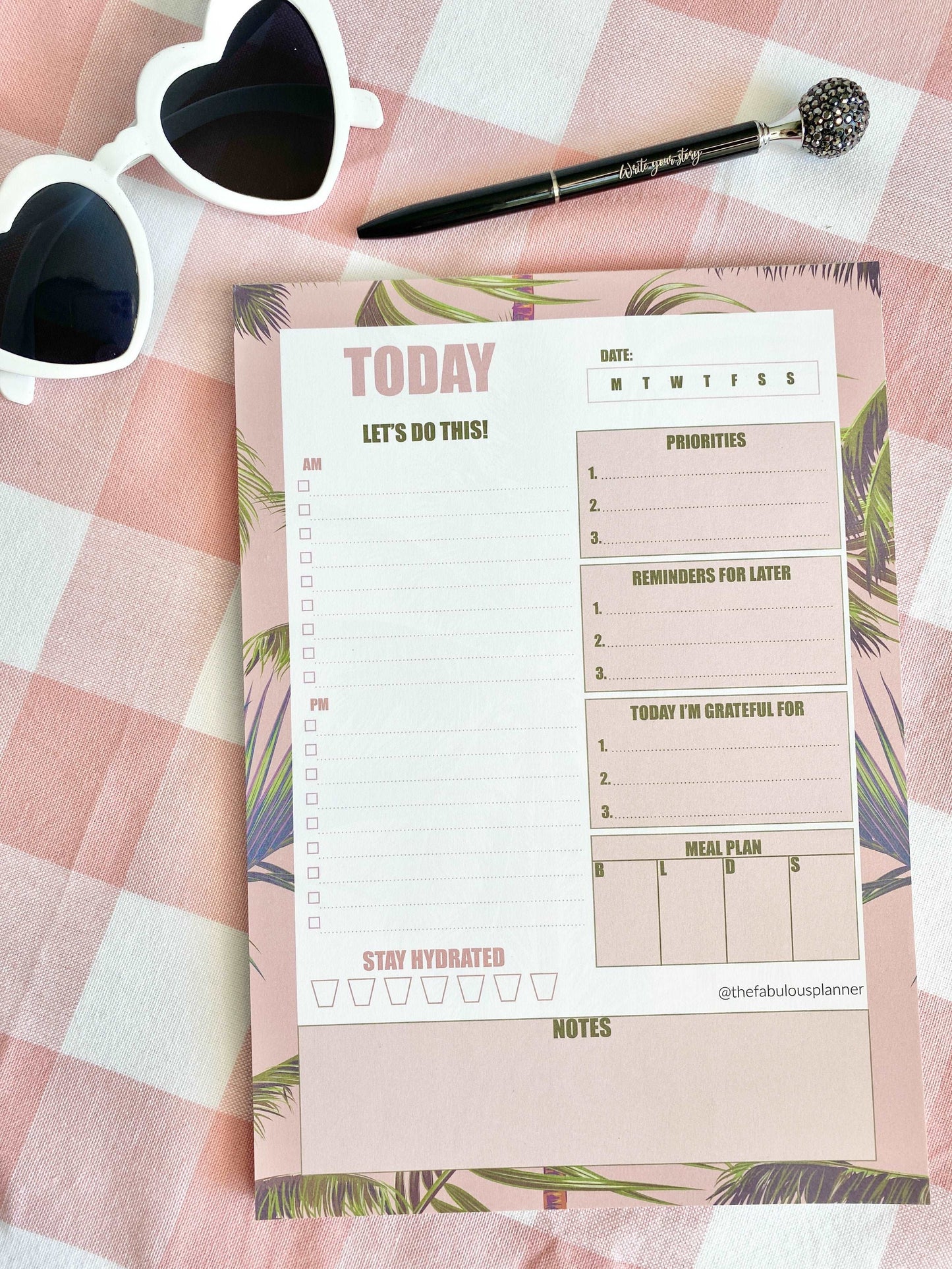 Pink tropical leaves 7x10 daily planner notepad styled on a pink gingham fabric background with sunglasses and a pen in soft natural light — for Carpe Diem Dream