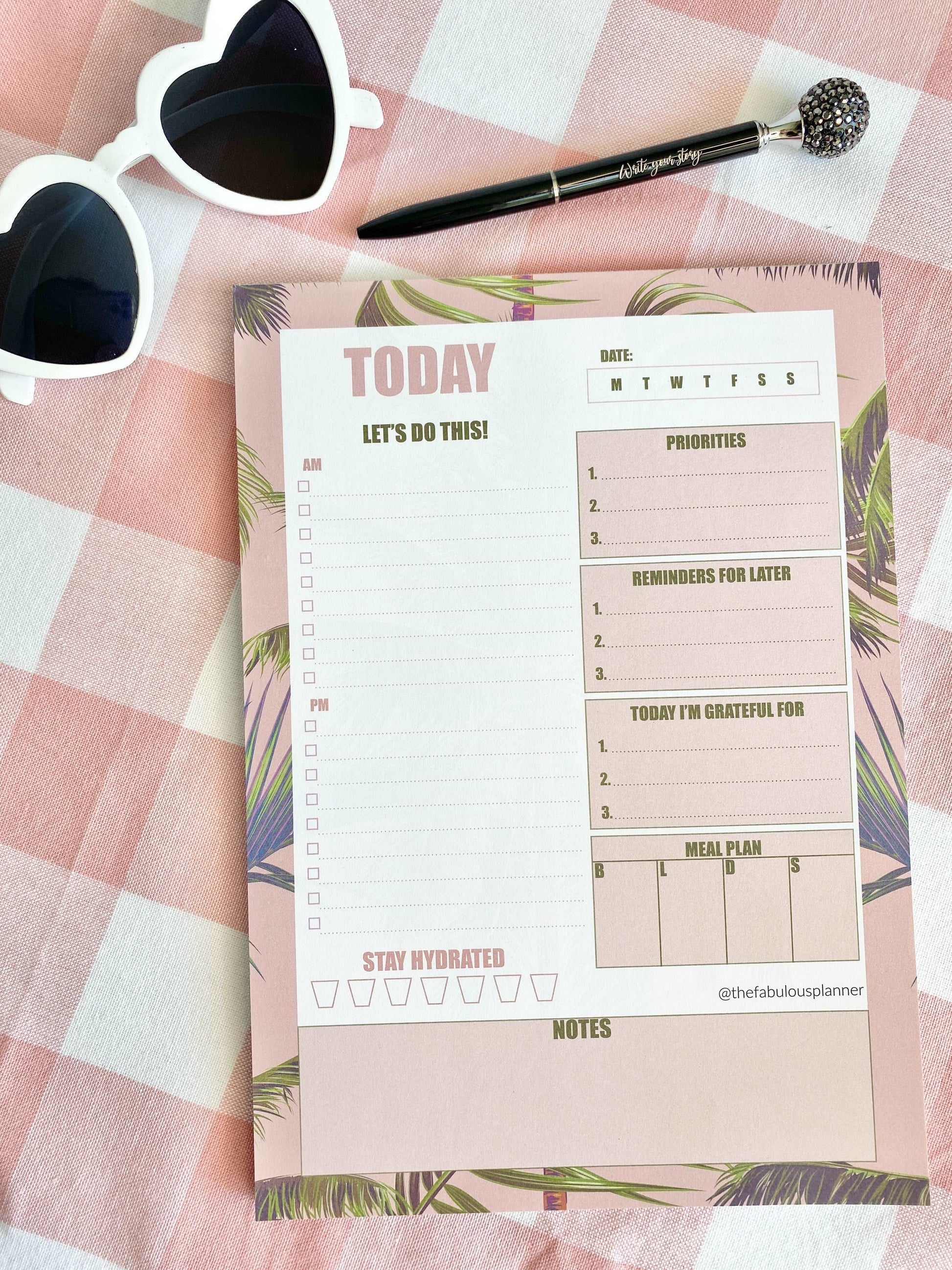 Pink tropical leaves 7x10 daily planner notepad styled on a pink gingham fabric background with sunglasses and a pen in soft natural light — for Carpe Diem Dream