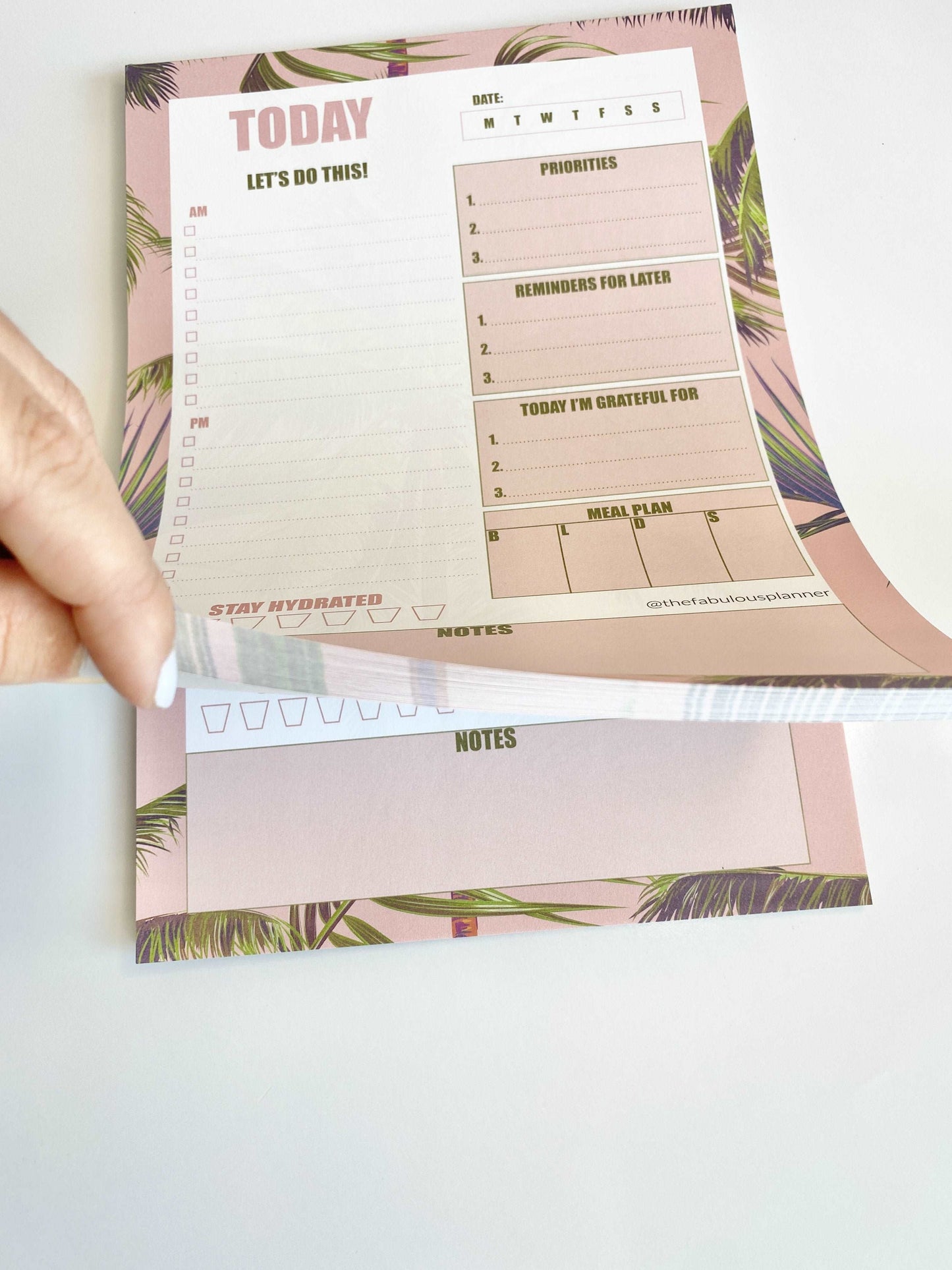 Pink tropical leaves 7x10 daily planner notepad with a hand lifting a page to show tear-off sheets on a white background — for Carpe Diem Dream