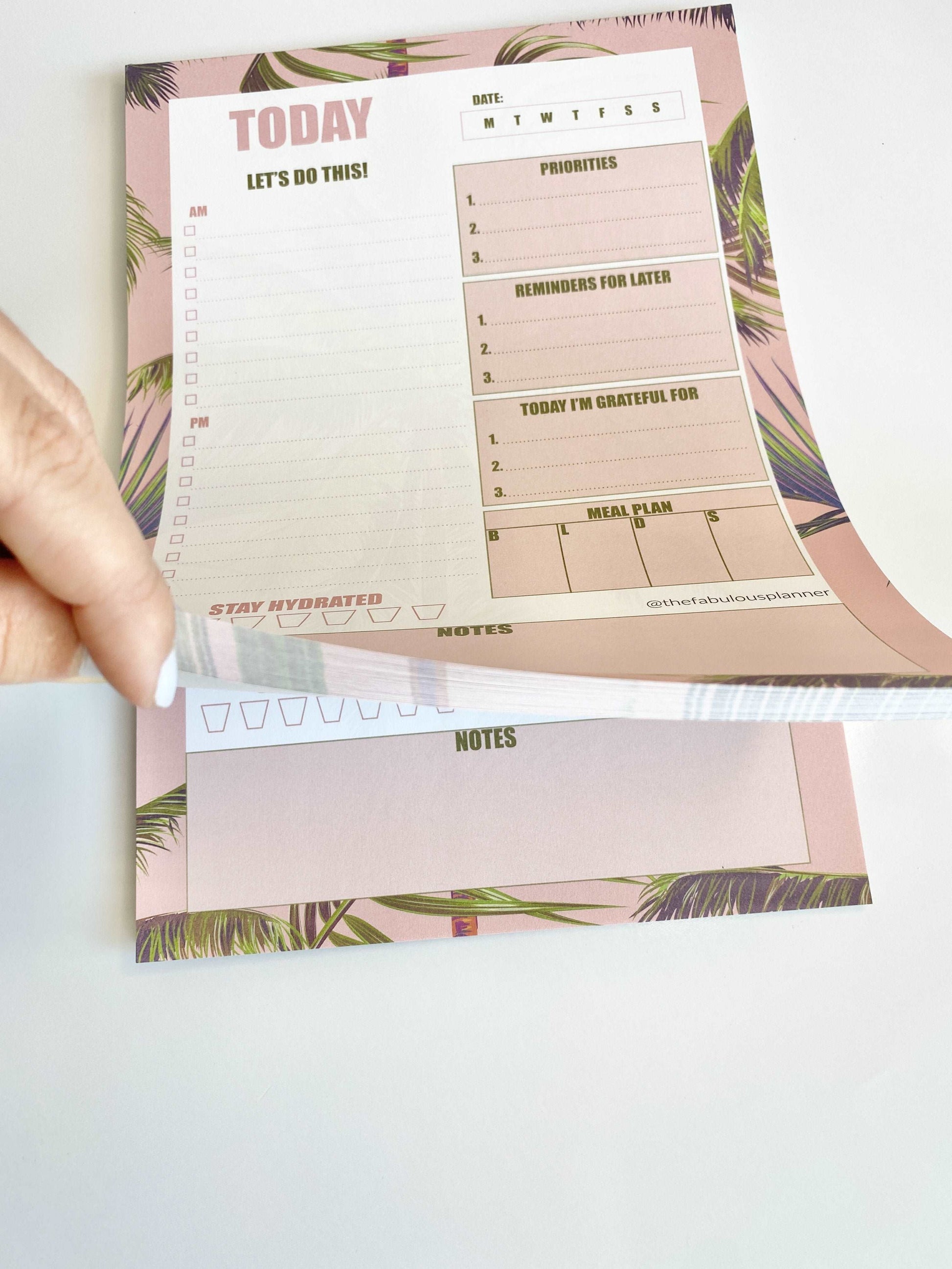 Pink tropical leaves 7x10 daily planner notepad with a hand lifting a page to show tear-off sheets on a white background — for Carpe Diem Dream