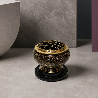 Close editorial view of a black-and-antique-gold brass resin incense burner highlighting the domed grid, pierced motifs, antiqued finish, ringed pedestal, and black wooden coaster. — for Carpe Diem Dream
