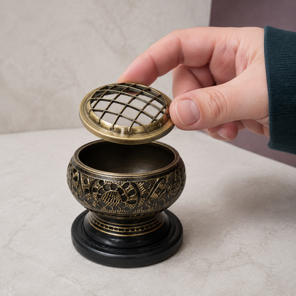 Compact black-and-antique-gold brass resin incense burner with a hand lifting the removable domed grid screen to reveal the empty bowl on a cool pale-stone surface. — for Carpe Diem Dream