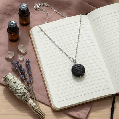 A round black lava stone essential oil necklace resting on an open journal beside small essential oil bottles, crystals, and dried lavender. — for Carpe Diem Dream