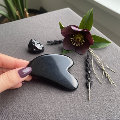 A hand holding a black obsidian gua sha facial tool on a textured surface with a flower and botanical accents nearby. — for Carpe Diem Dream