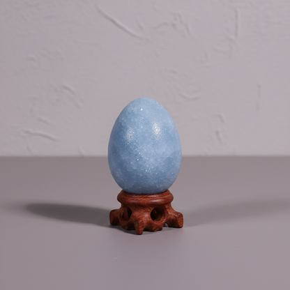 A polished blue celestite crystal egg on a carved wooden stand displayed against a neutral textured wall with soft studio lighting and minimal styling. — for Carpe Diem Dream