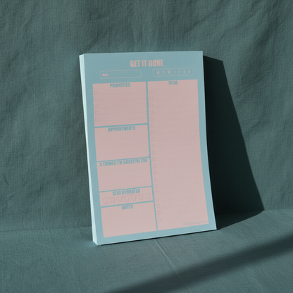 Blue and pink 7x10 daily planner notepad displayed upright on a muted teal textured background with soft natural shadow — for Carpe Diem Dream