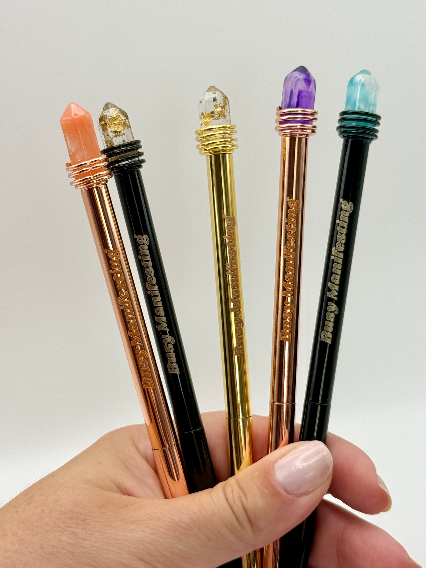 Hand holding metallic Busy Manifesting crystal mystic pens with faux crystal tops against white background — for Carpe Diem Dream