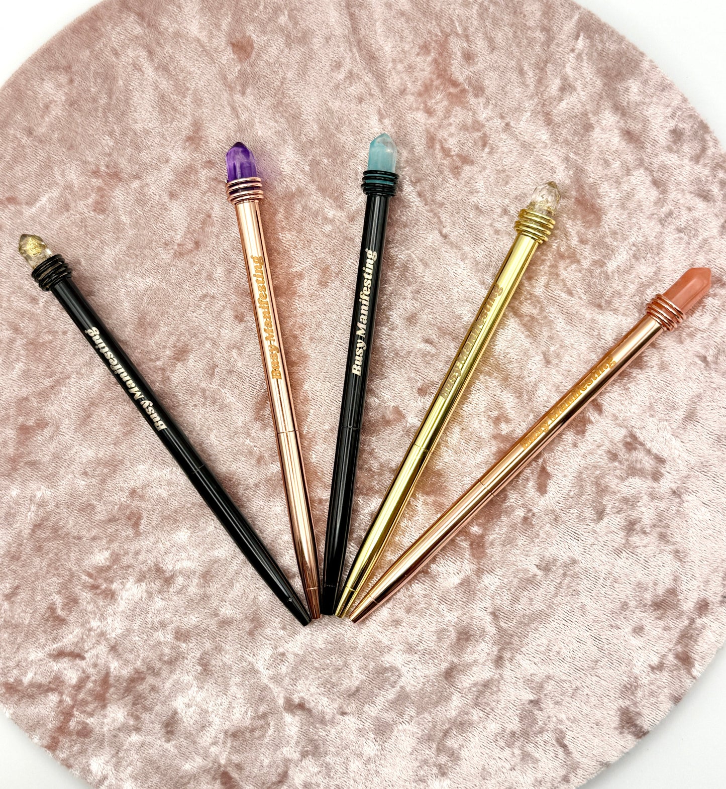 Set of metallic Busy Manifesting crystal mystic pens with faux crystal tops displayed in a fan arrangement on textured blush background — for Carpe Diem Dream