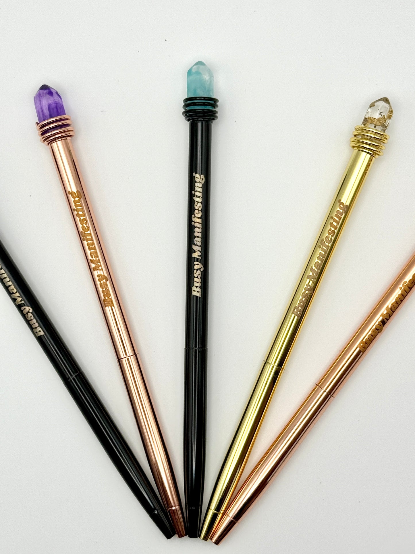 Set of metallic Busy Manifesting crystal mystic pens with faux crystal tops arranged in a fan layout on white background — for Carpe Diem Dream