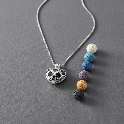 A silver heart-shaped cage diffuser necklace displayed on a gray surface with six interchangeable lava stone beads arranged beside it. — for Carpe Diem Dream