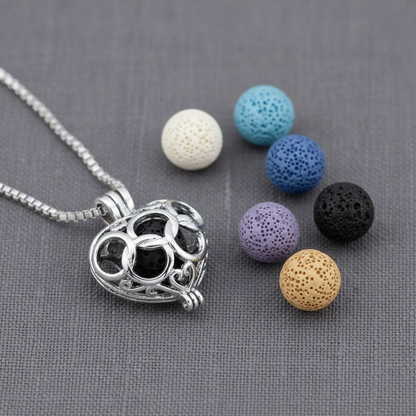 A silver heart-shaped cage diffuser necklace displayed with six interchangeable lava stone beads in assorted colors on a textured gray background. — for Carpe Diem Dream