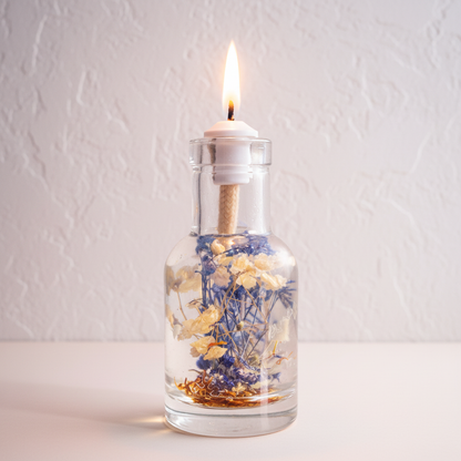 A Cancer zodiac oil candle in a clear glass vessel with embedded white and blue botanicals is displayed on a soft neutral surface against a textured light wall with a steady flame. — for Carpe Diem Dream