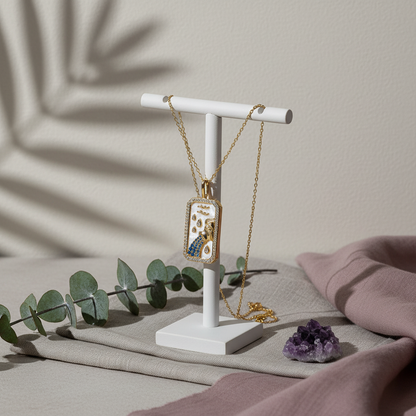 An 18K gold zodiac pendant necklace displayed on a white jewelry stand with eucalyptus, mauve fabric, and an amethyst crystal in soft natural light. — for Carpe Diem Dream