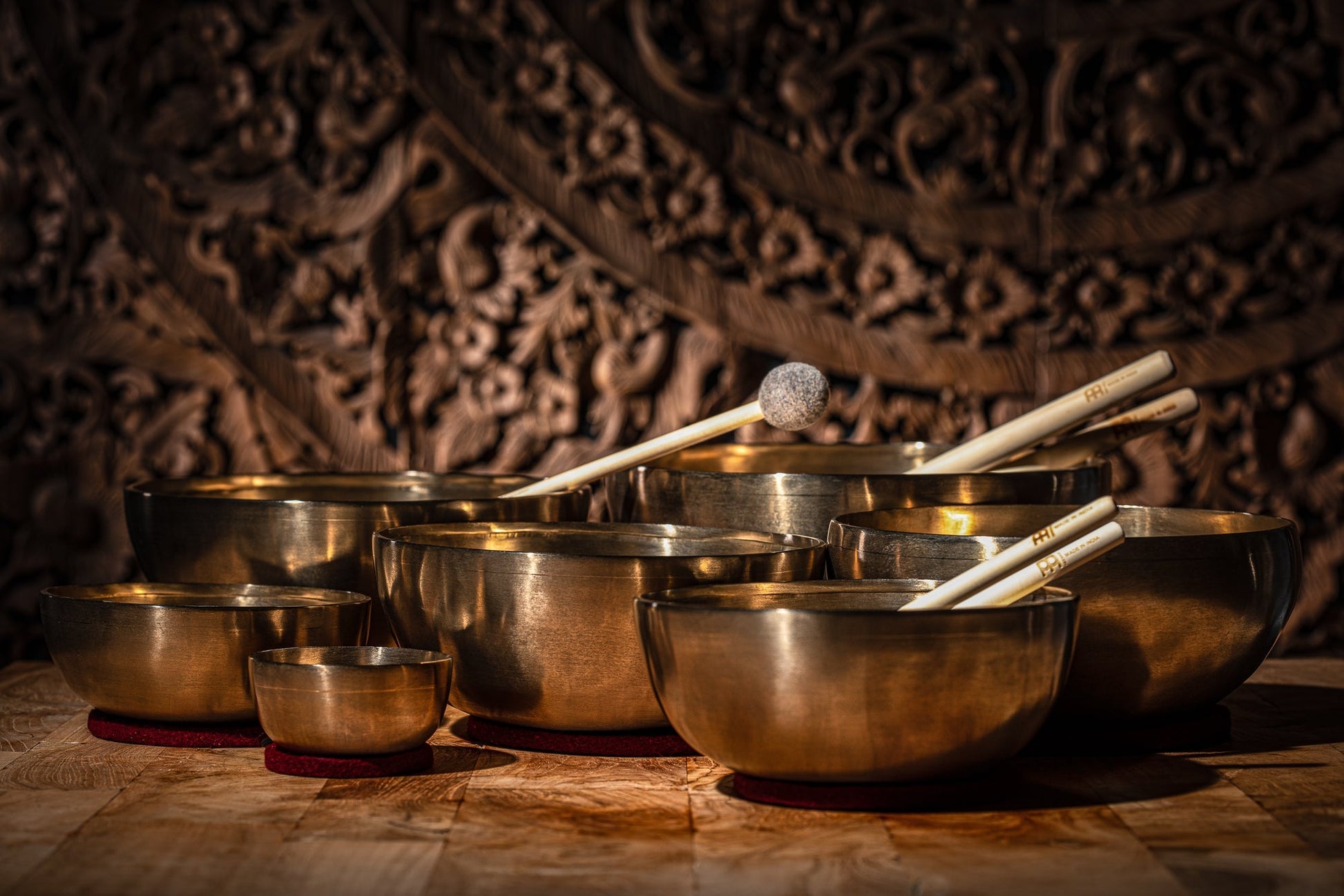 Seven handcrafted bronze singing bowls with wooden-handled mallets resting inside, displayed on red felt rings against a carved wooden backdrop. — for Carpe Diem Dream