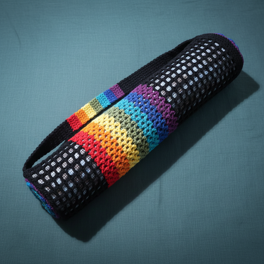 A rolled black crochet chakra yoga mat bag with rainbow stripes is displayed on a dark teal-grey textured linen background with cool diffused studio lighting. — for Carpe Diem Dream