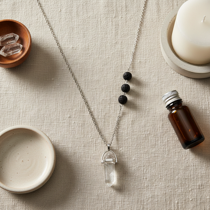 A clear crystal pendant necklace with black lava stone beads displayed on a textured linen surface beside an amber essential oil bottle and ceramic bowls. — for Carpe Diem Dream