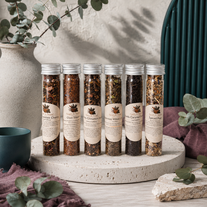 Cozy Winter Tea Sampler tubes displayed in the Carpe Diem Dream botanical studio with eucalyptus, mauve linen, natural stone, and dark teal accents. — for Carpe Diem Dream