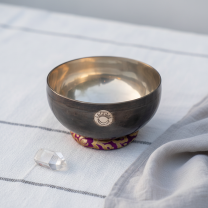 Hand-hammered Crown Chakra New Moon singing bowl tuned to B note resting on a purple cushion on soft linen with a clear quartz crystal in a minimal studio setting. — for Carpe Diem Dream