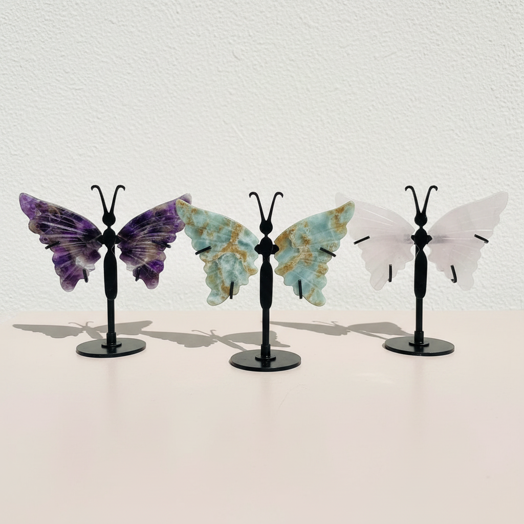 Three crystal butterfly wing variants—amethyst, amazonite, and rose quartz—displayed on black stands in a clean, minimal setting to show color and size comparison. — for Carpe Diem Dream