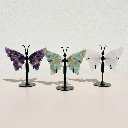 Three crystal butterfly wing variants—amethyst, amazonite, and rose quartz—displayed on black stands in a clean, minimal setting to show color and size comparison. — for Carpe Diem Dream