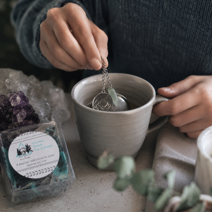 Crystal Charm Tea Infuser – authentic tea infuser shown in a candid preparation moment with hands placing the metal mesh strainer and crystal charm into a neutral ceramic mug, styled with clear packaging, natural stone, cool-toned crystals, soft linen, and cool diffused daylight. — for Carpe Diem Dream