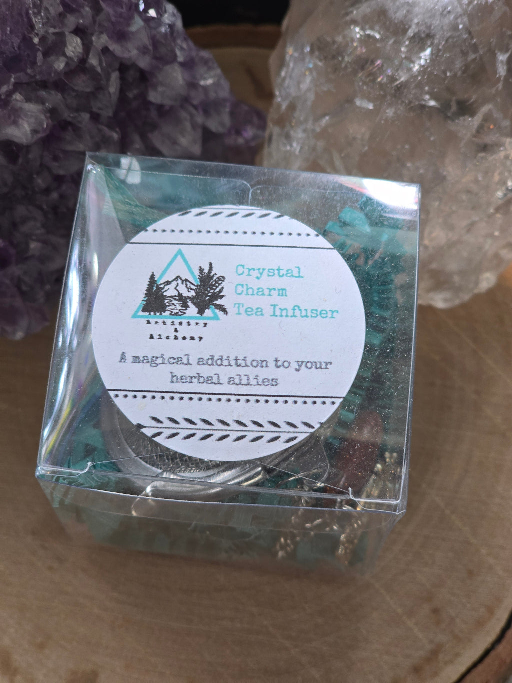 Crystal Charm Tea Infuser – Authentic supplier image showing the packaged tea infuser with crystal charms in a clear display box, styled in front of amethyst and clear quartz crystals on a natural wood surface. — for Carpe Diem Dream