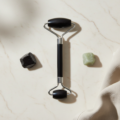 A black crystal face roller placed on a light marble surface beside two small polished stones with soft natural shadows. — for Carpe Diem Dream