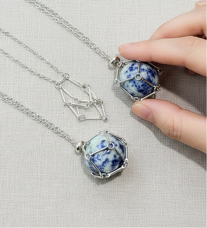 A crystal gem holder necklace with a blue stone in a silver wire cage held by a hand on a neutral textured background. — for Carpe Diem Dream
