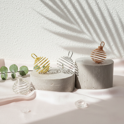 A set of silver, gold, and copper spiral crystal cage pendants arranged on stone pedestals with soft botanical shadows on a neutral background. — for Carpe Diem Dream