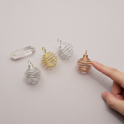 A set of silver, gold, and copper spiral crystal cage pendants shown beside a hand on a soft neutral surface to demonstrate size. — for Carpe Diem Dream
