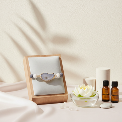 A crystal and gold lava stone stretch bracelet displayed on a wooden stand beside amber essential oil bottles, a floating flower bowl, and soft neutral decor. — for Carpe Diem Dream