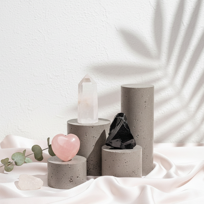 Clear quartz point, pink rose quartz heart, and raw black obsidian magnet arranged on concrete cylinders with soft silk draping and botanical shadows on a textured wall. — for Carpe Diem Dream