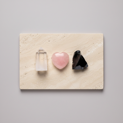 Clear quartz point, rose quartz heart, and raw black obsidian magnet arranged on a travertine slab against a soft neutral backdrop. — for Carpe Diem Dream