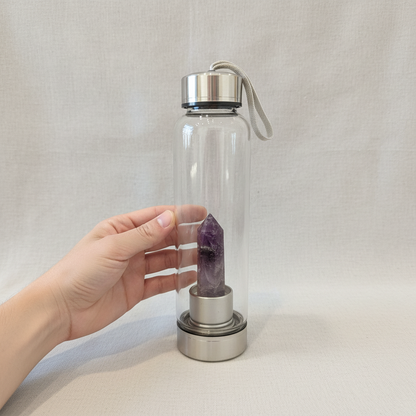 A crystal infused glass water bottle with an amethyst point shown beside a hand for size reference against a neutral fabric background. — for Carpe Diem Dream