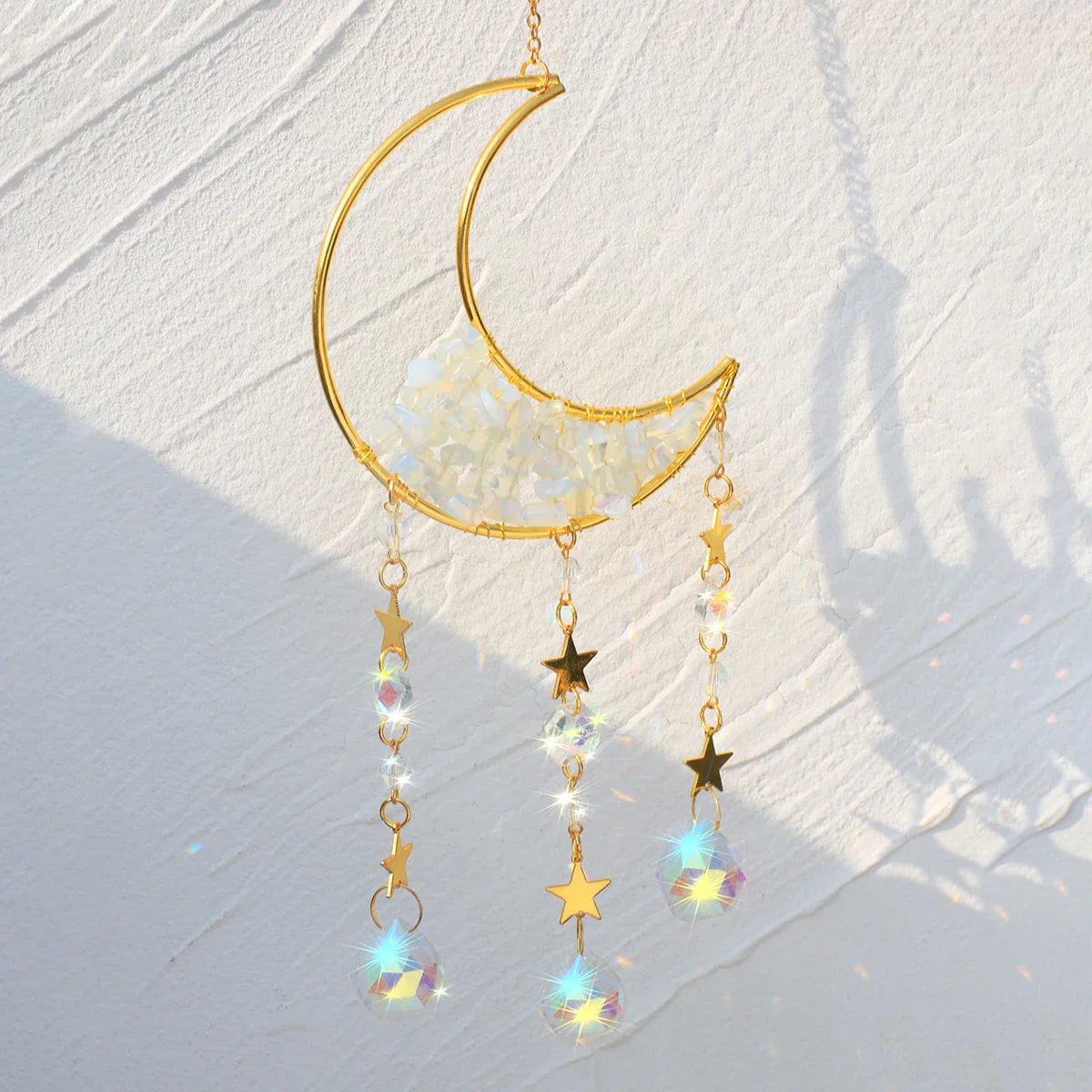 A clear quartz crystal moon suncatcher with a gold crescent frame, translucent stone chips, and hanging prism drops displayed in bright natural sunlight casting vivid rainbow reflections and soft shadows. — for Carpe Diem Dream